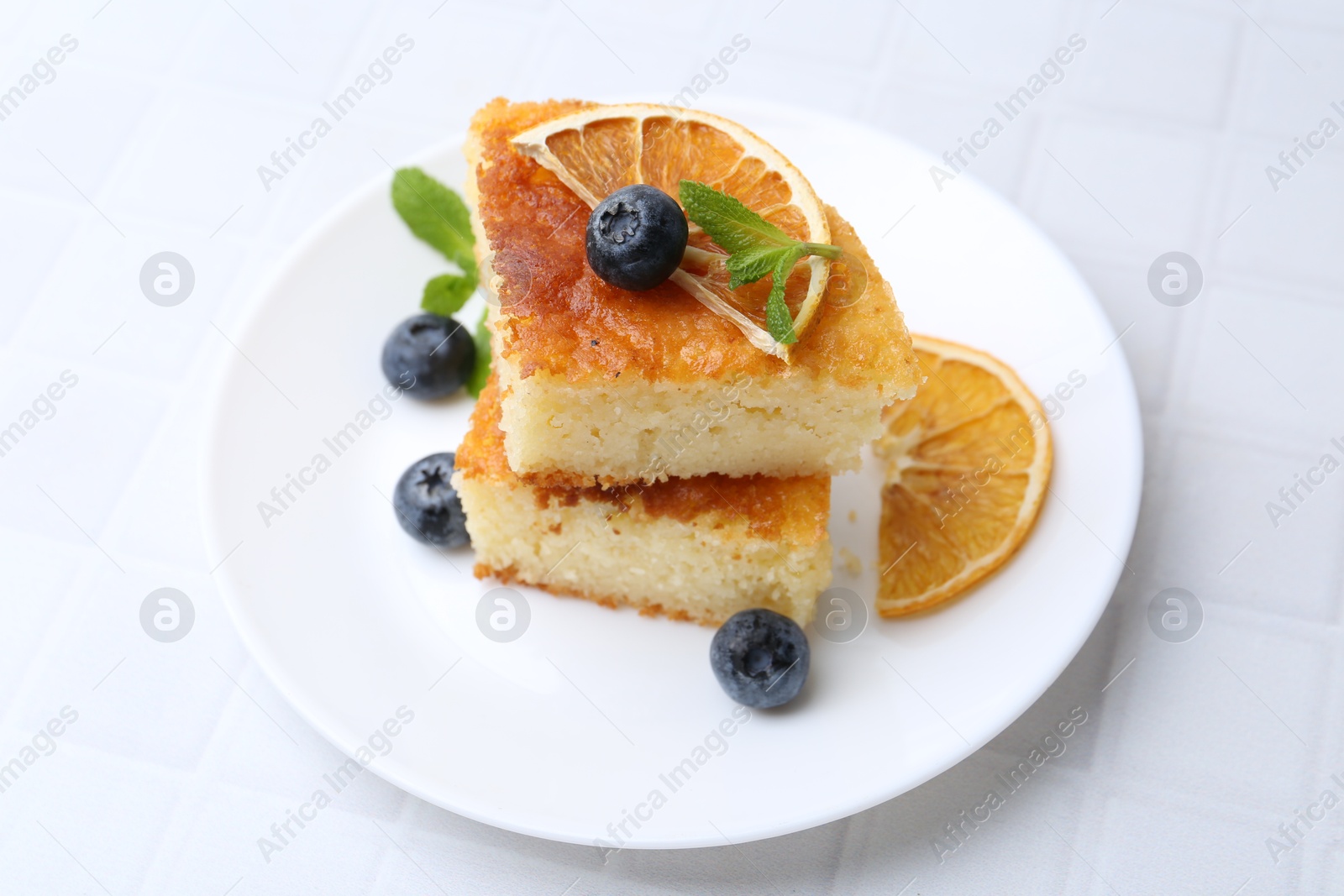 Photo of Pieces of delicious semolina cake with blueberries and orange slices on white tiled table, closeup