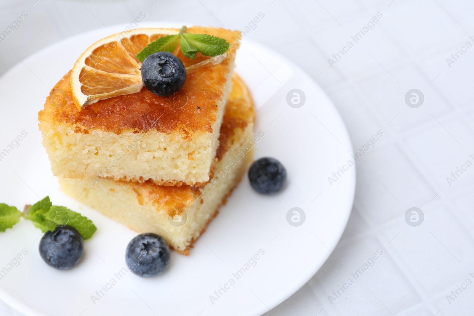 Photo of Pieces of delicious semolina cake with blueberries and orange slices on white tiled table, closeup