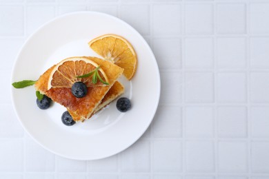 Photo of Pieces of delicious semolina cake with blueberries and orange slices on white tiled table, top view. Space for text