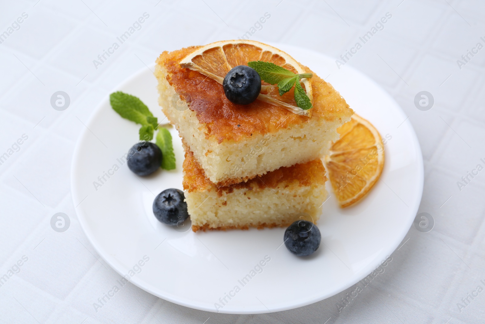 Photo of Pieces of delicious semolina cake with blueberries and orange slices on white tiled table, closeup