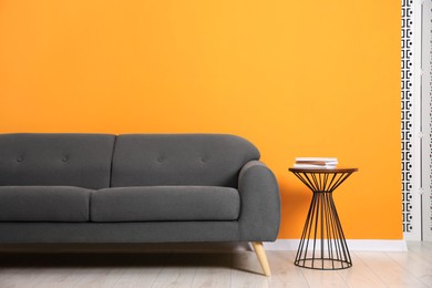 Photo of Stylish sofa and side table with books near orange wall indoors