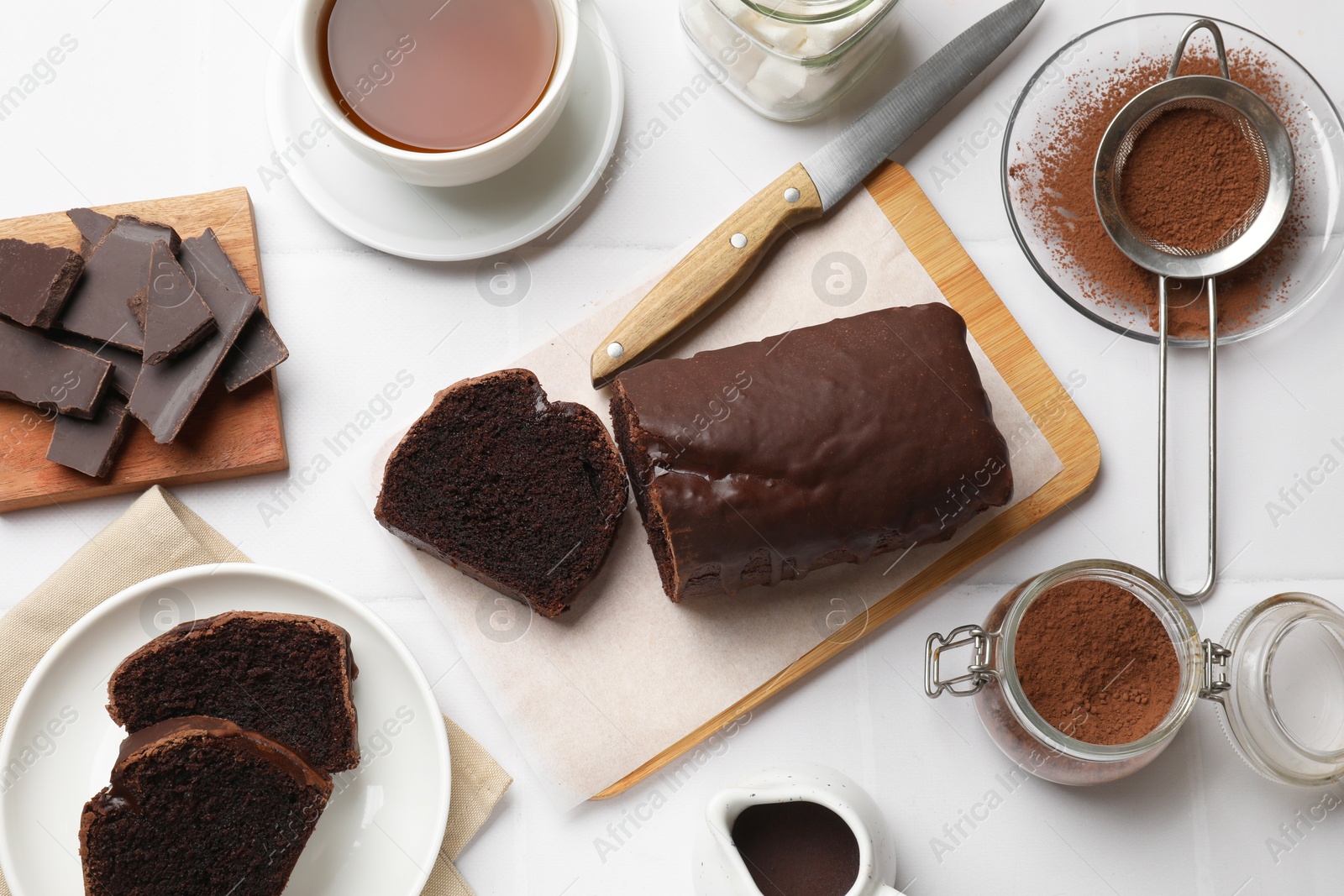 Photo of Delicious cut chocolate sponge cake and ingredients on white tiled table, flat lay