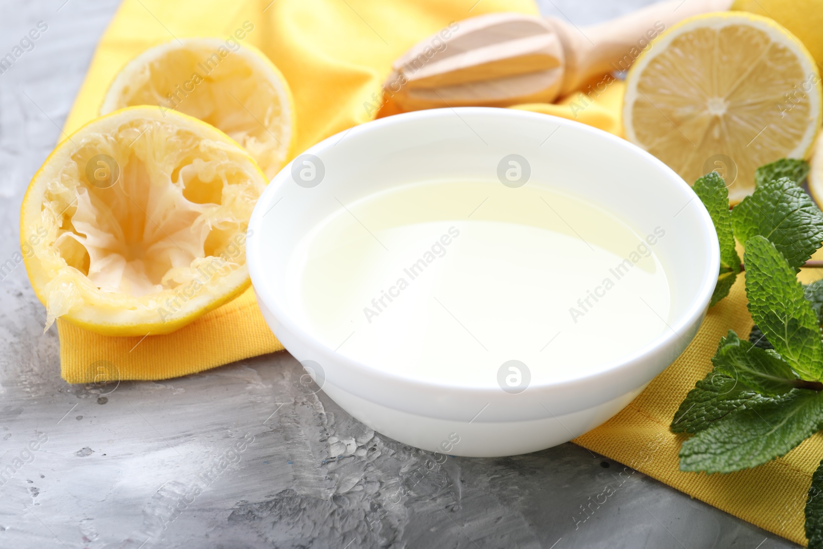 Photo of Fresh lemon juice in bowl, fruits, mint and squeezer on grey table, closeup