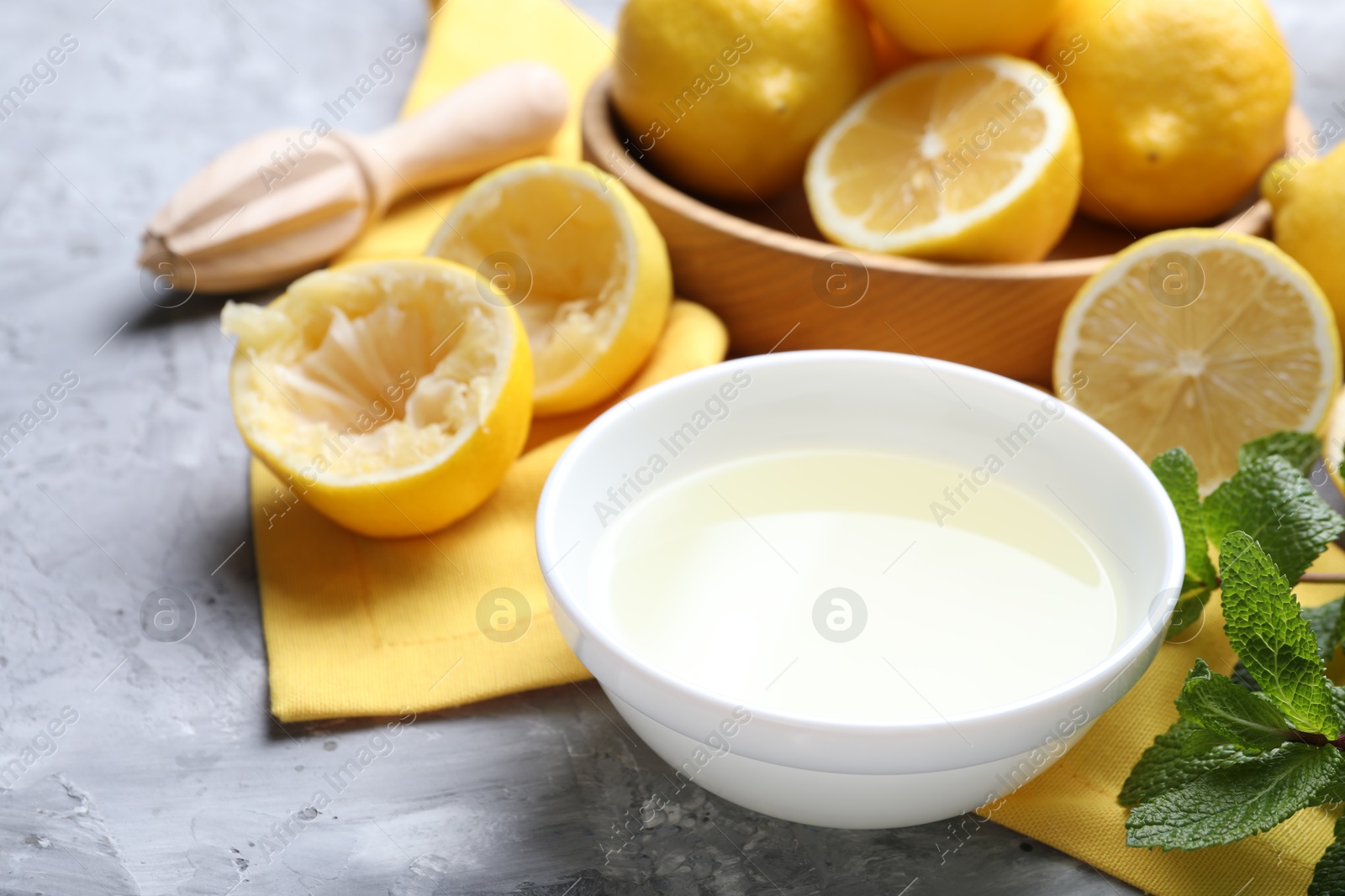 Photo of Fresh lemon juice in bowl, fruits, mint and squeezer on grey table, closeup