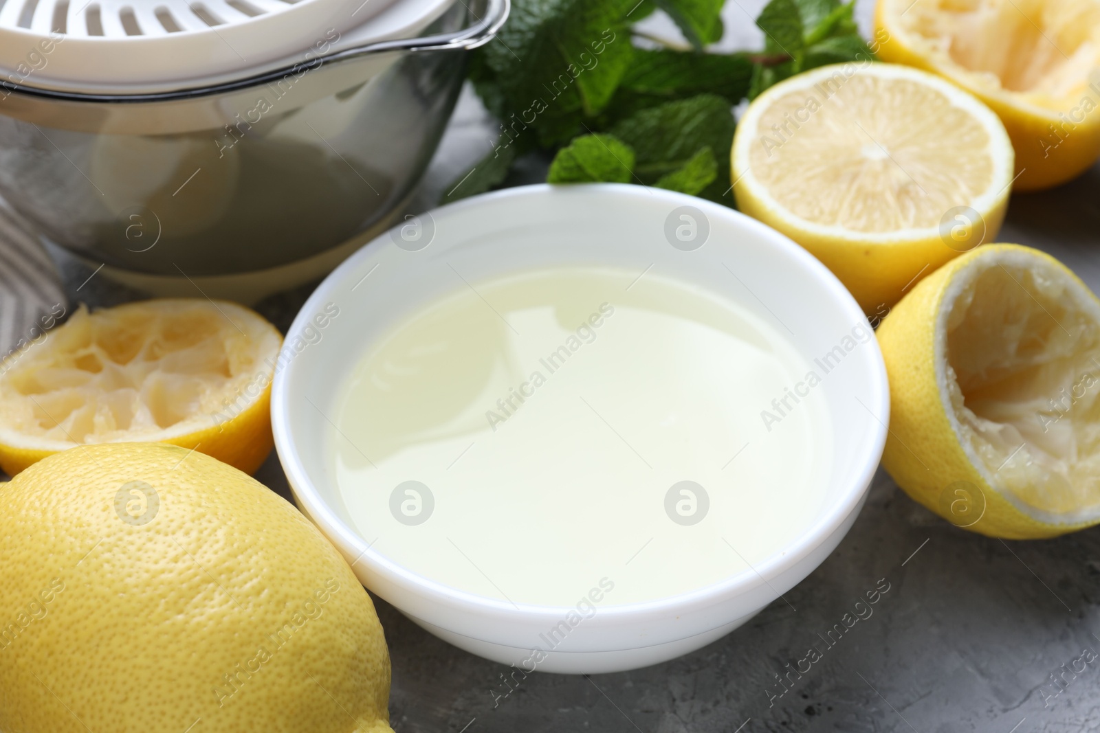 Photo of Fresh lemon juice in bowl, fruits, mint and juicer on grey table, closeup