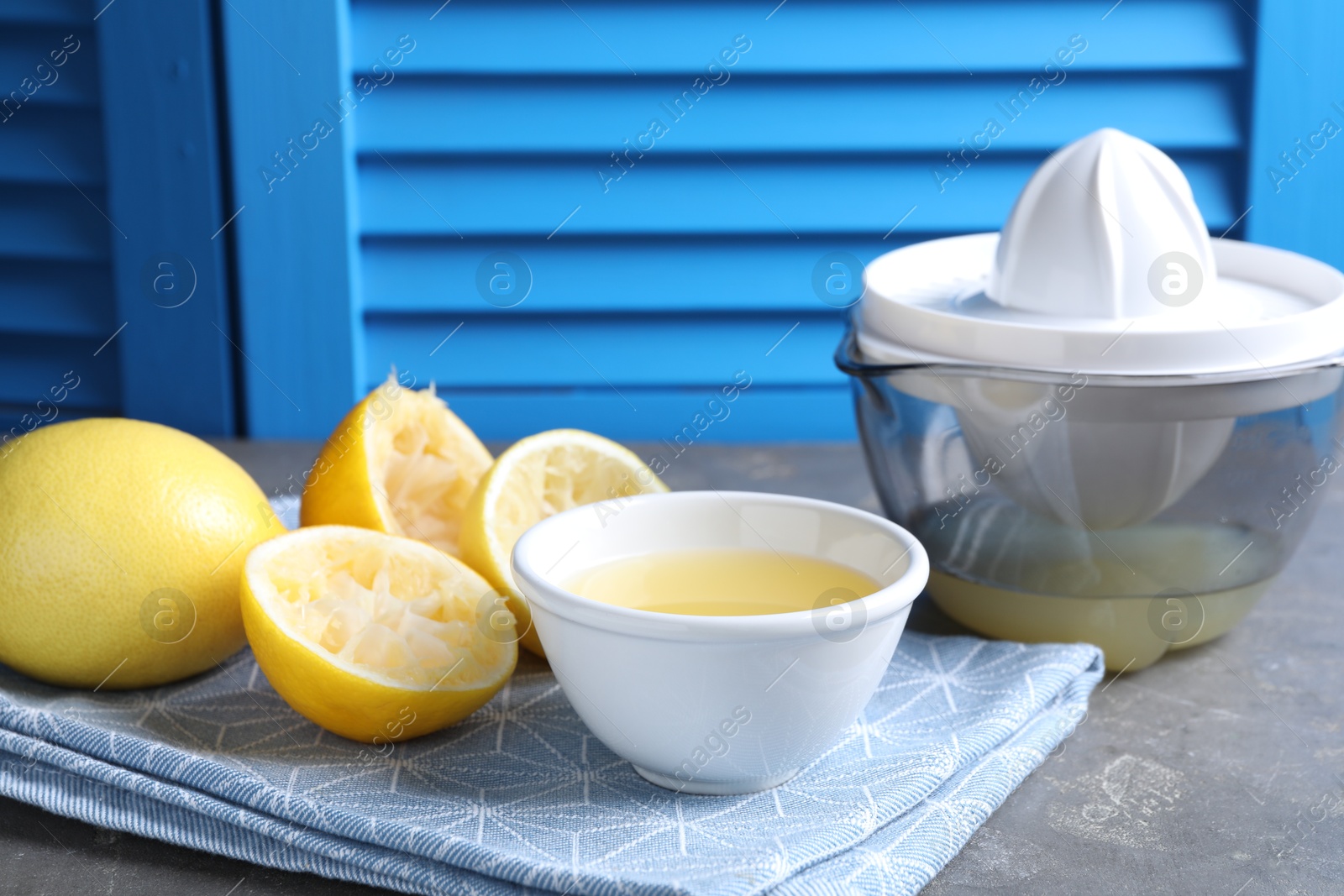 Photo of Fresh lemon juice in bowl, fruits and juicer on grey table