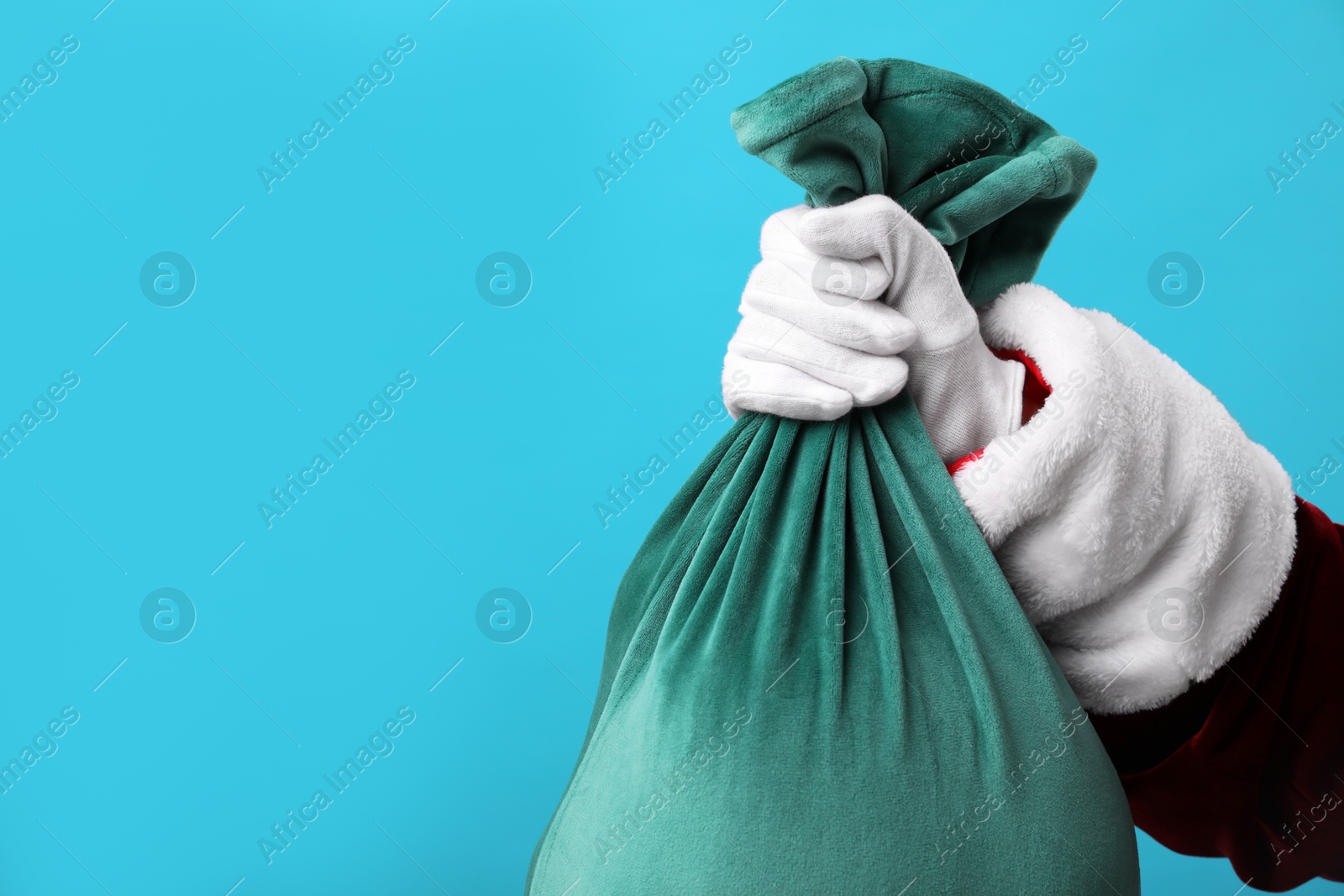 Photo of Santa Claus with green bag of Christmas gifts on light blue background, closeup. Space for text