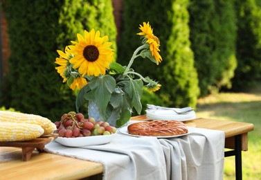 Photo of Vase with sunflowers, grapes, pie and corncobs on wooden table in garden