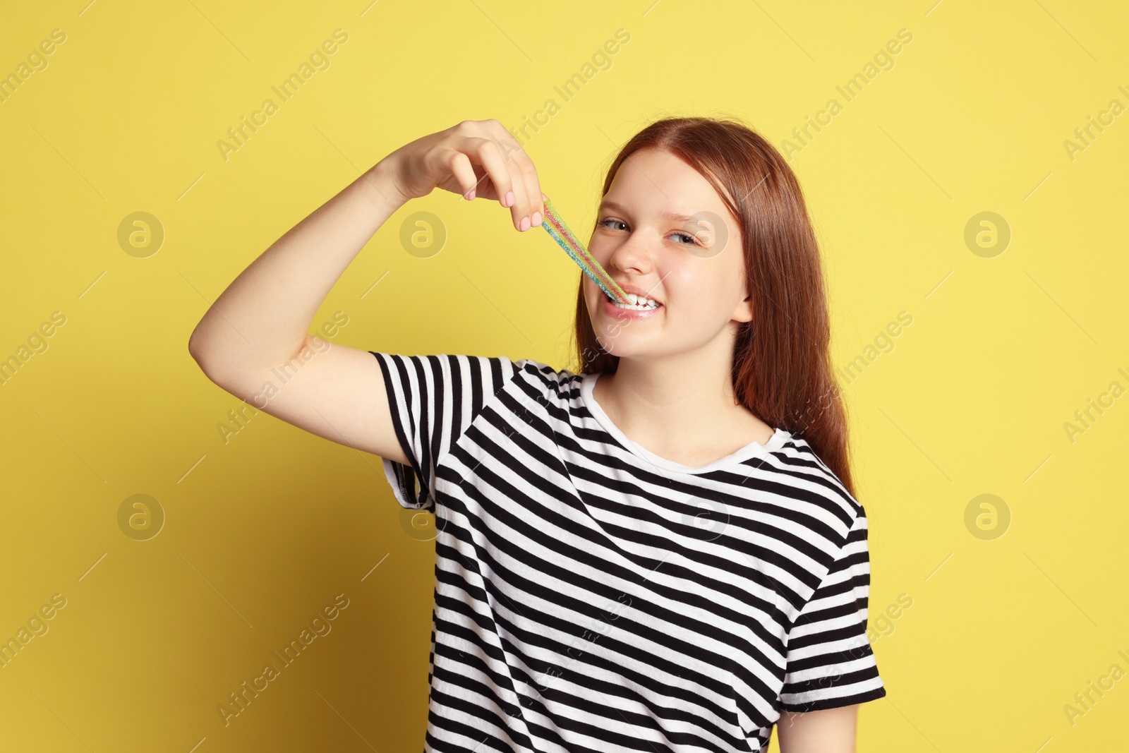 Photo of Teenage girl eating tasty rainbow sour belt on yellow background