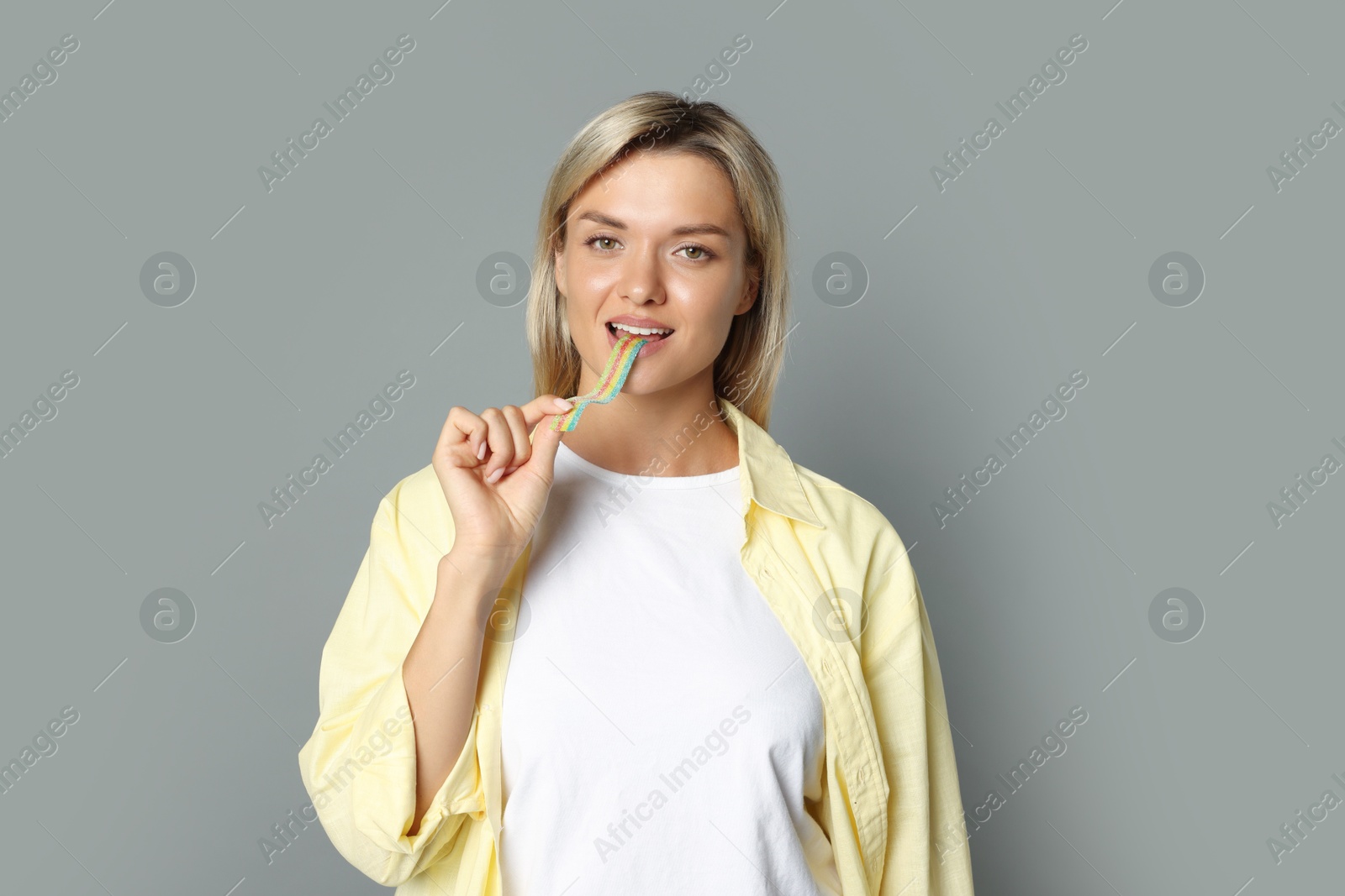 Photo of Young woman eating tasty rainbow sour belt on grey background