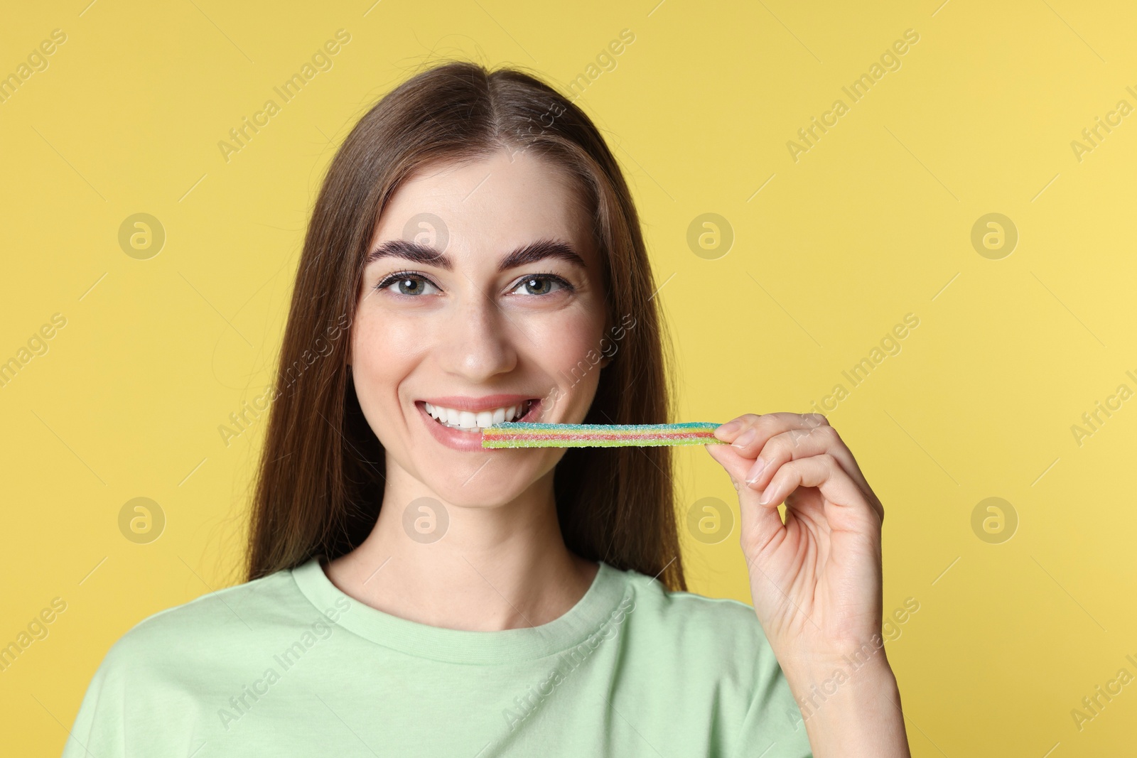 Photo of Young woman eating tasty rainbow sour belt on yellow background