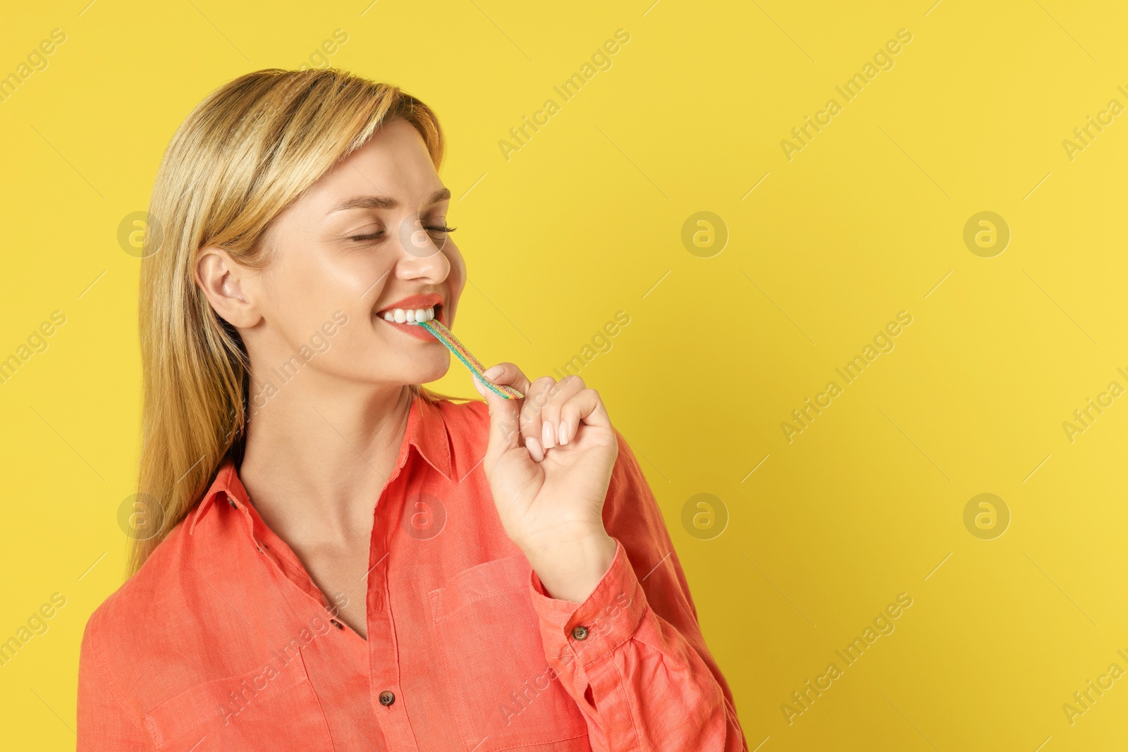 Photo of Young woman eating tasty rainbow sour belt on yellow background, space for text