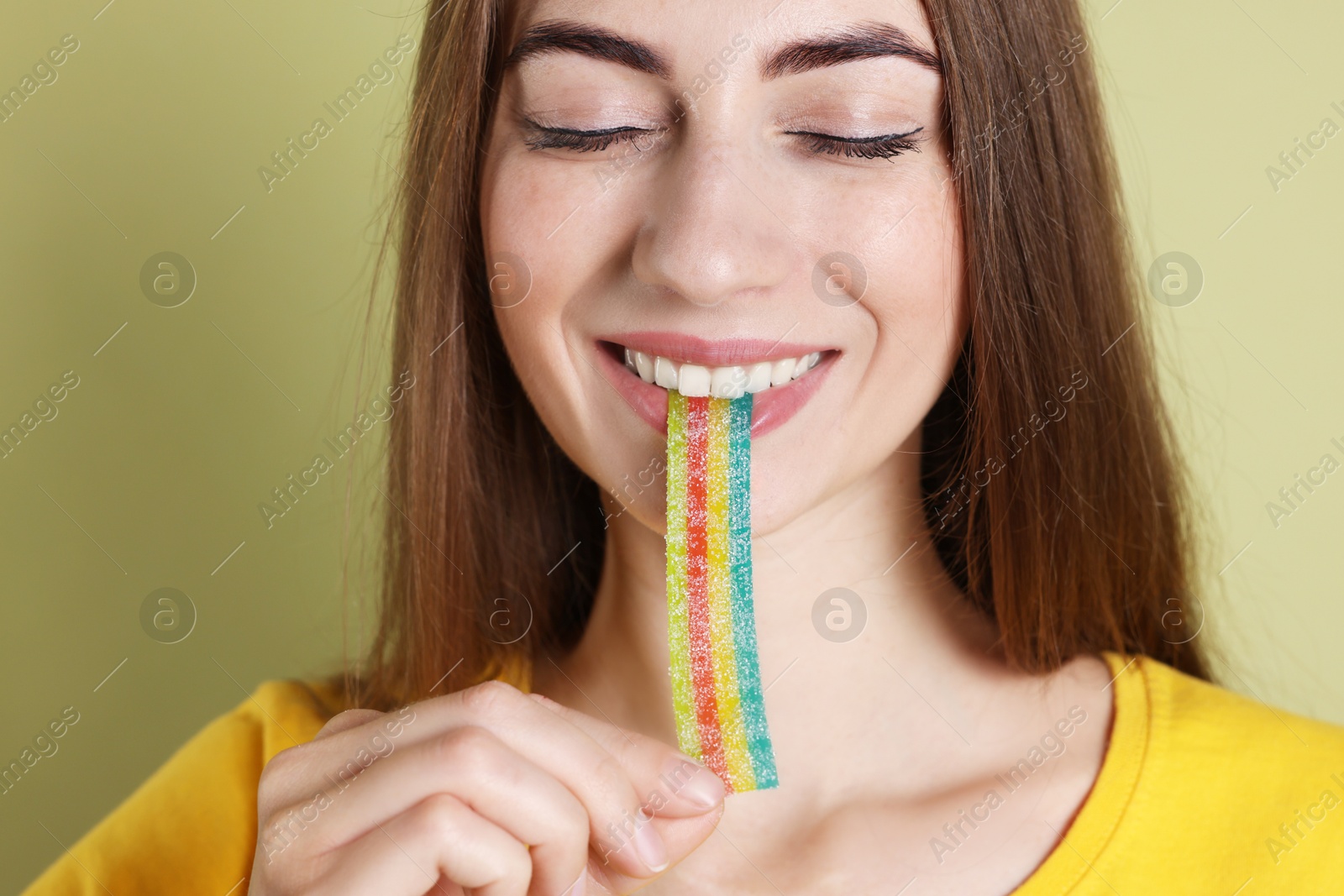 Photo of Young woman eating tasty rainbow sour belt on olive background, closeup