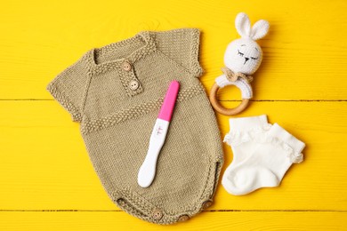 Photo of Pregnancy test, baby onesie, toy and socks on yellow wooden table, flat lay