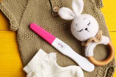 Photo of Pregnancy test, baby onesie, toy and socks on yellow wooden table, flat lay