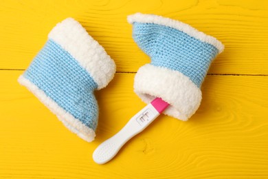 Photo of Pregnancy test and baby shoes on yellow wooden table, flat lay