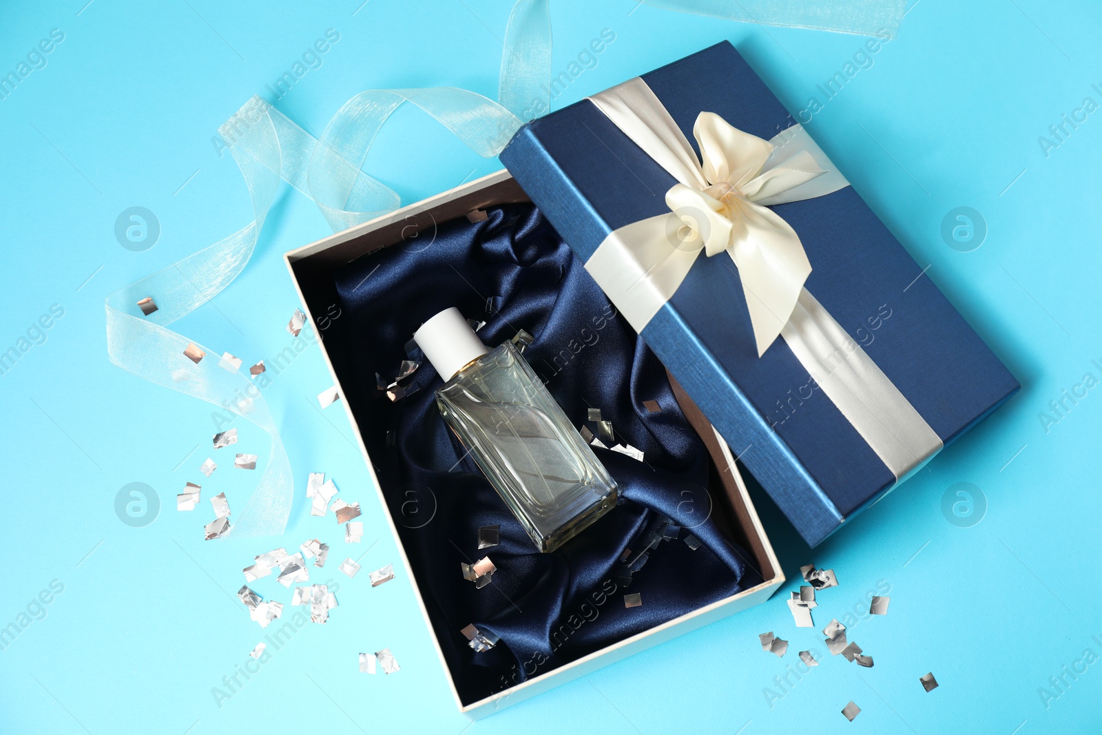 Photo of Perfume bottle in gift box and confetti on light blue background, top view