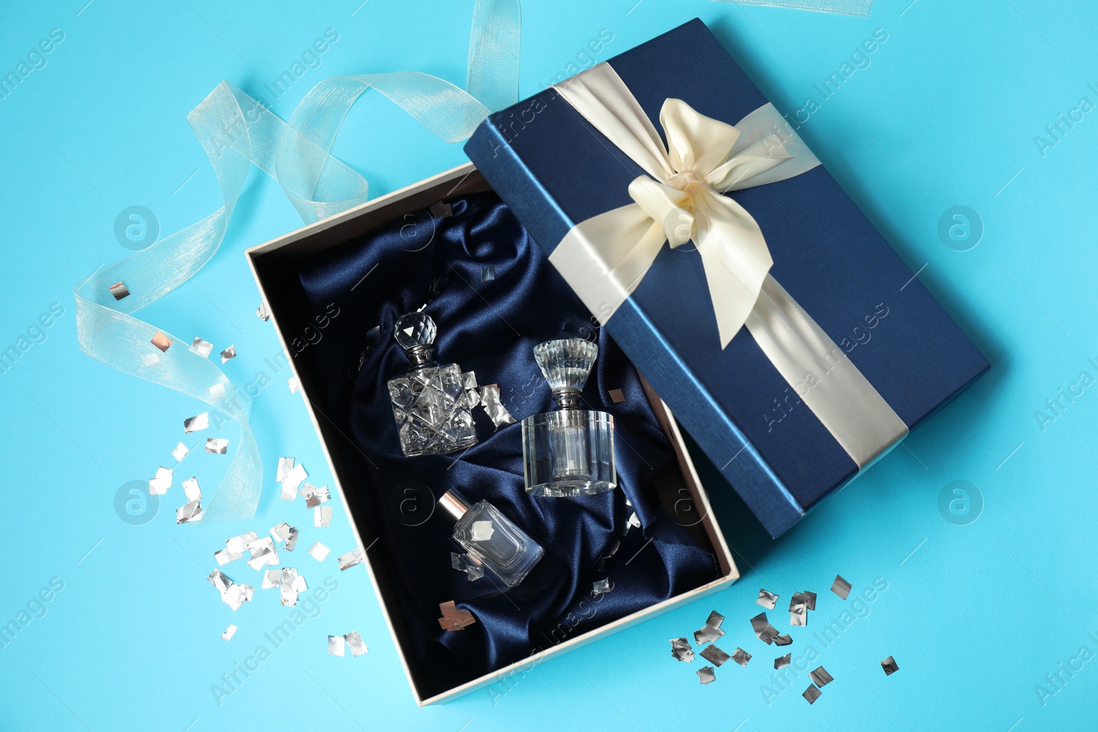 Photo of Perfume bottles in gift box and confetti on light blue background, top view