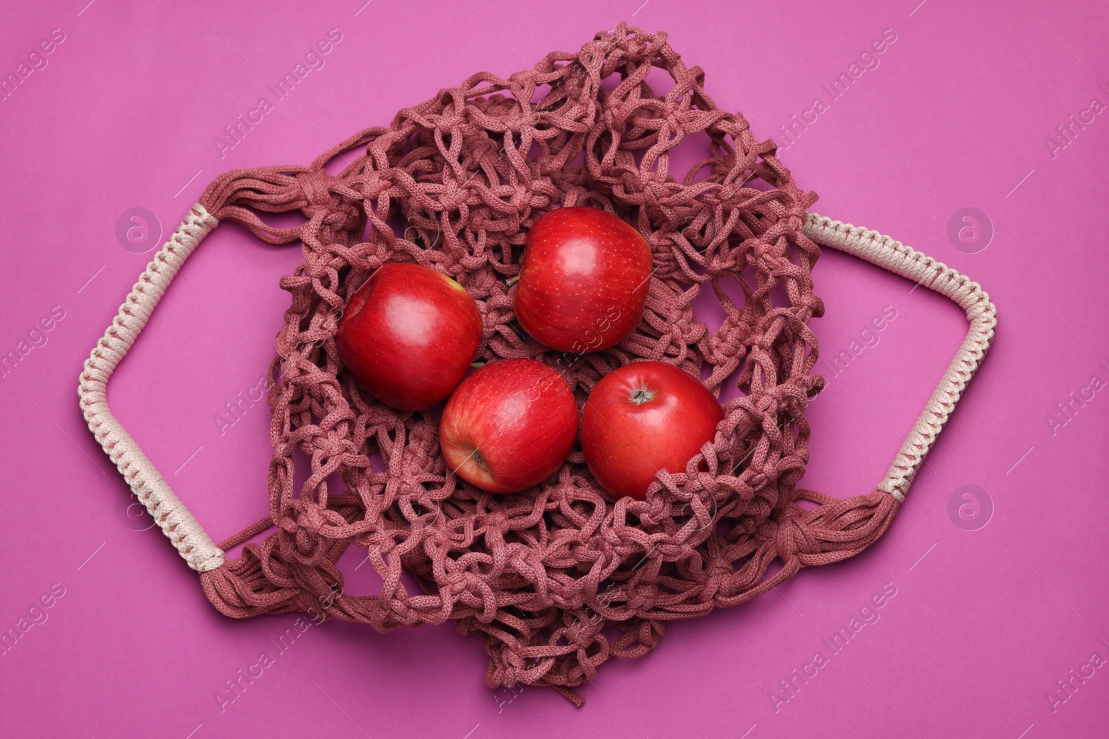 Photo of Handmade macrame shopping bag with apples on crimson background, top view