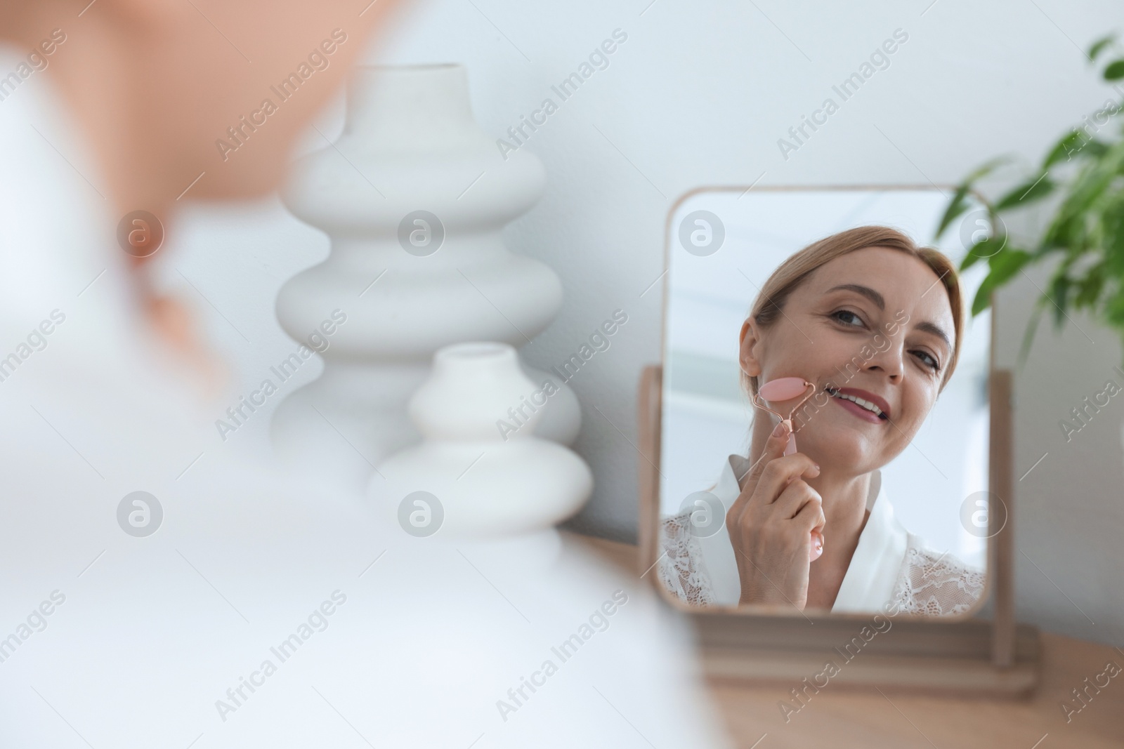 Photo of Smiling woman doing facial self massage with roller near mirror at home