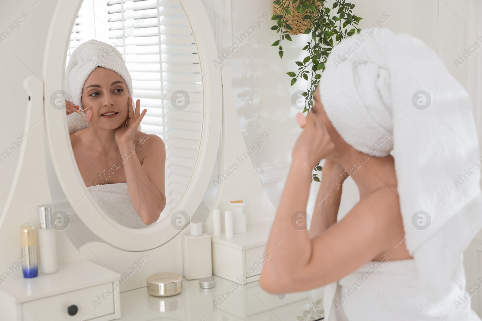 Photo of Smiling woman doing facial self massage with roller near mirror in bathroom