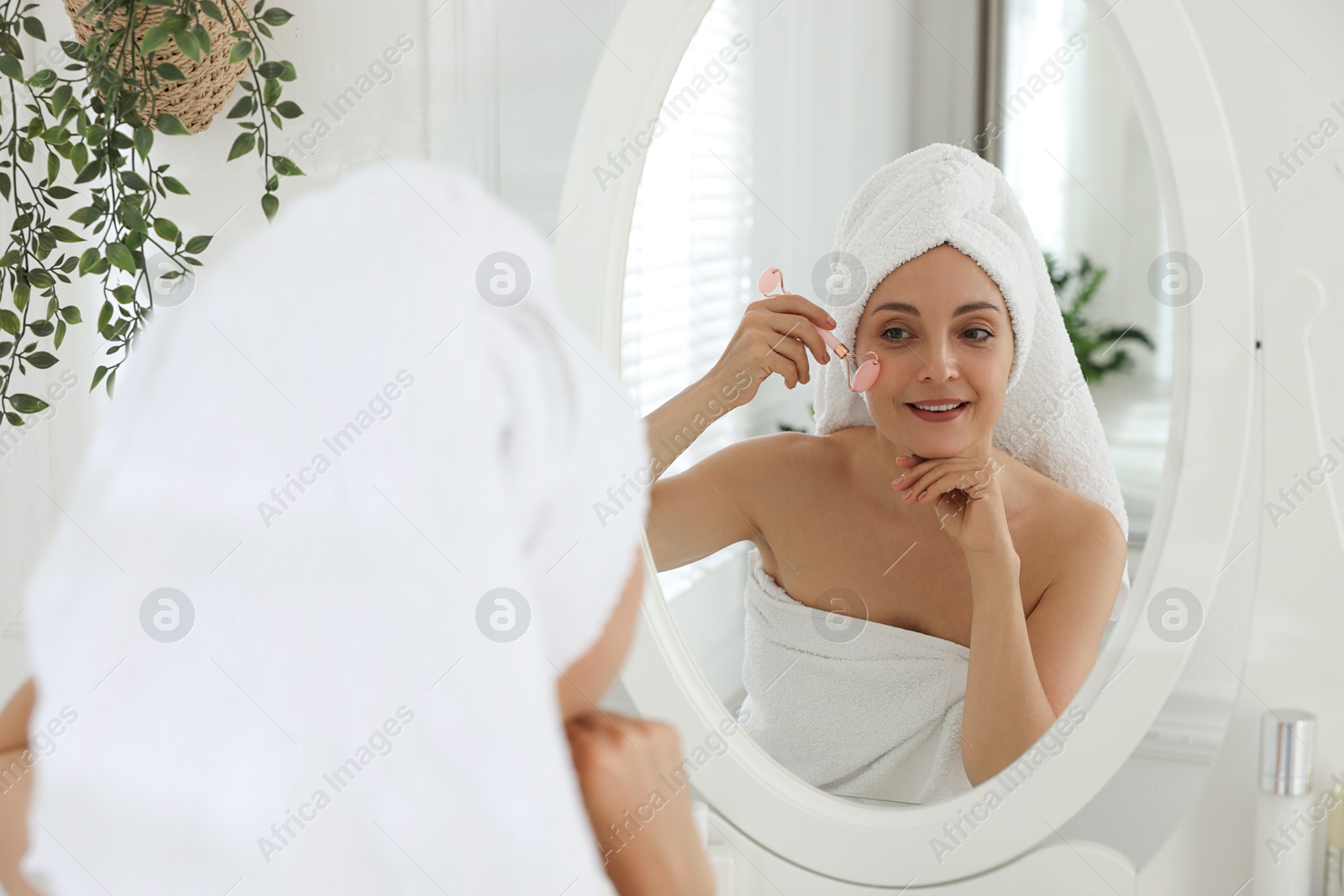 Photo of Smiling woman doing facial self massage with roller near mirror in bathroom