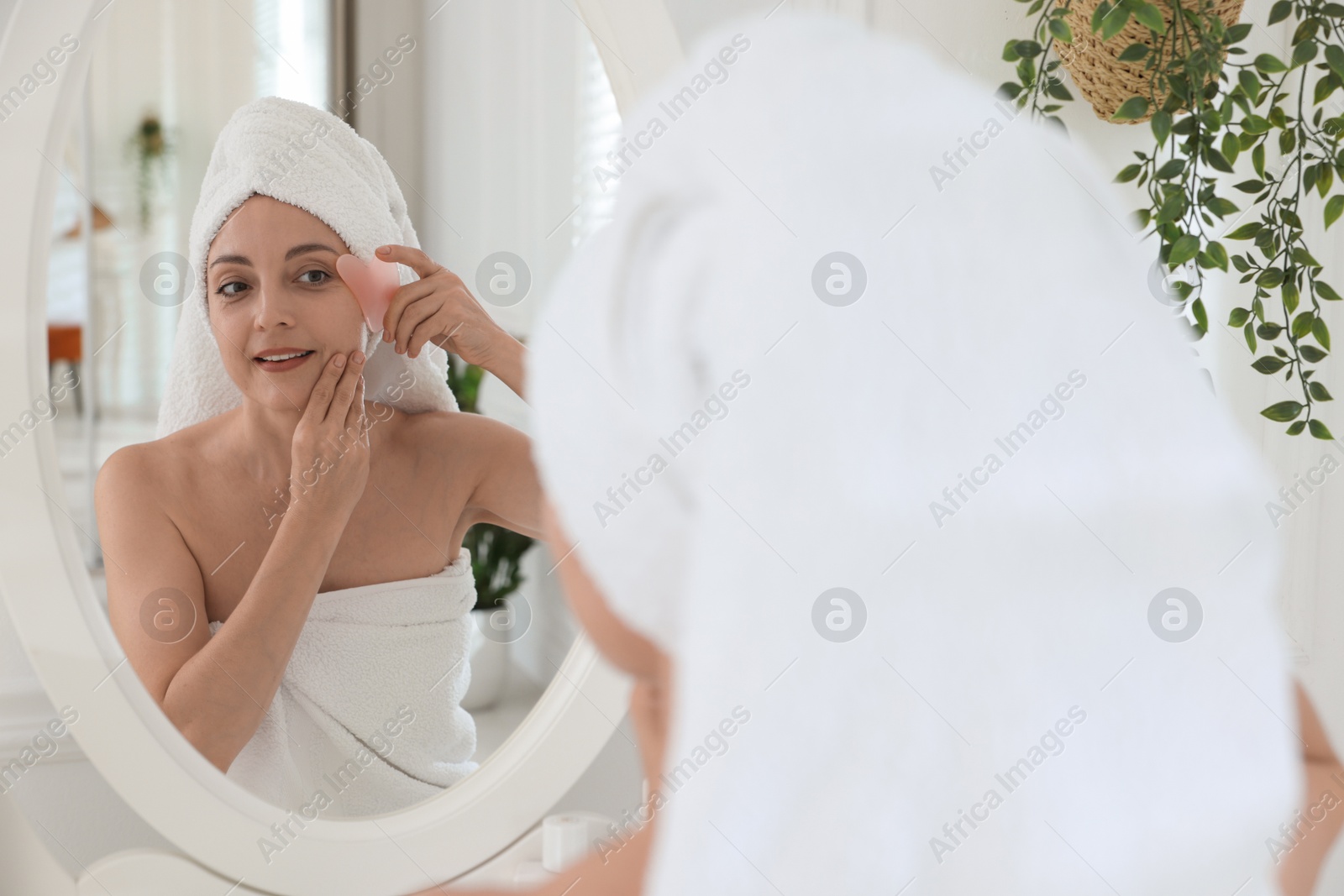 Photo of Smiling woman doing facial self massage with gua sha tool near mirror in bathroom