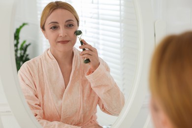 Photo of Beautiful woman doing facial self massage with roller near mirror in bathroom