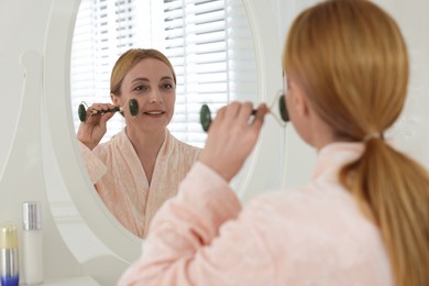 Photo of Smiling woman doing facial self massage with roller near mirror in bathroom