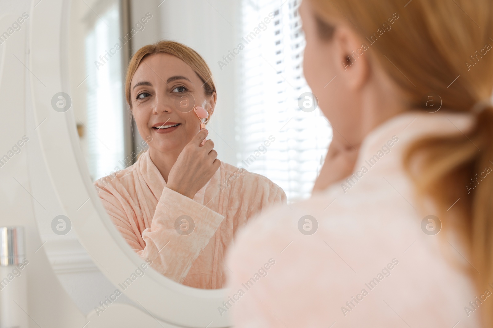 Photo of Smiling woman doing facial self massage with roller near mirror in bathroom