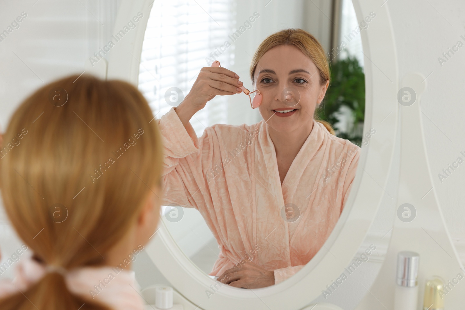 Photo of Smiling woman doing facial self massage with roller near mirror in bathroom