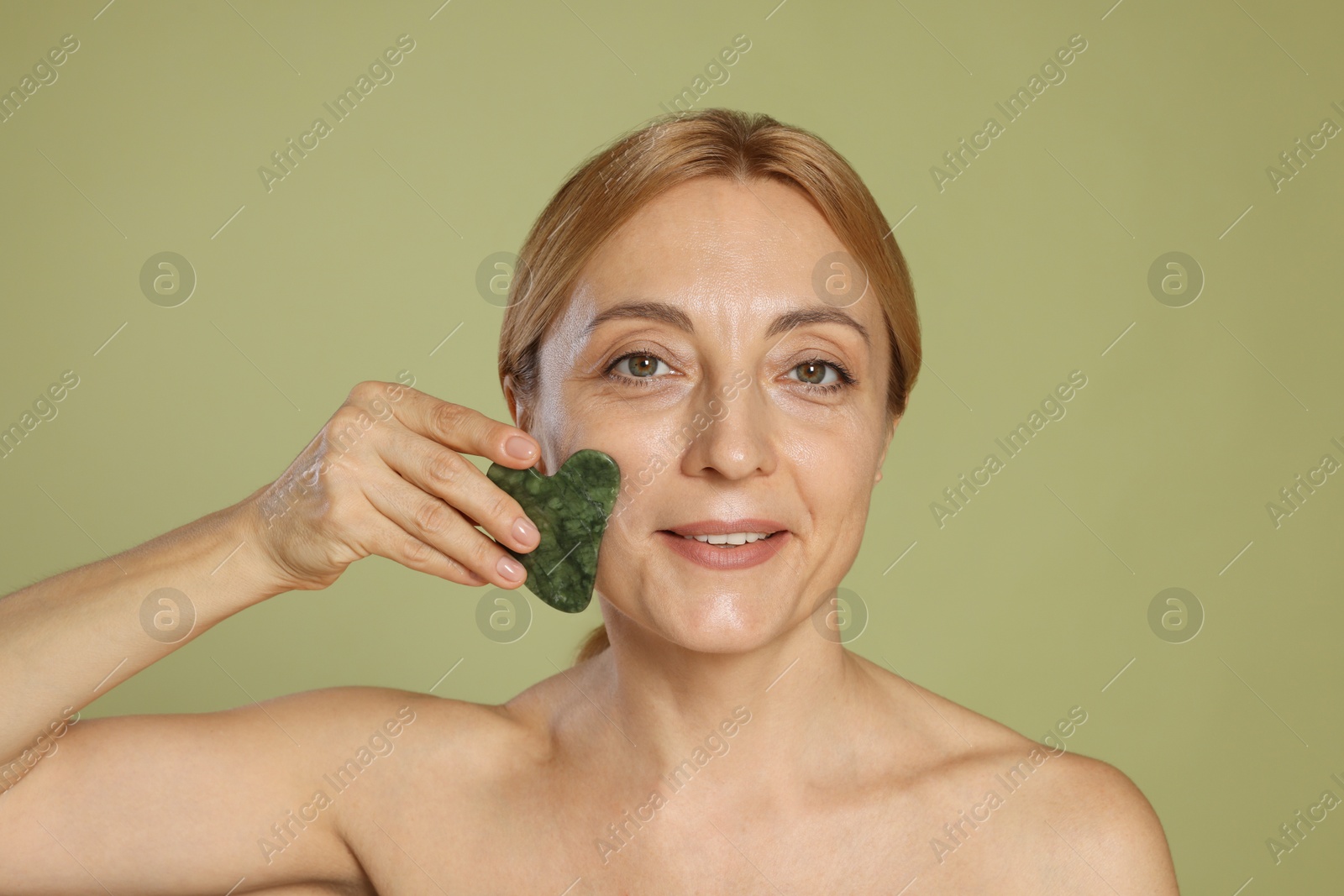 Photo of Smiling woman doing facial self massage with gua sha tool on green background