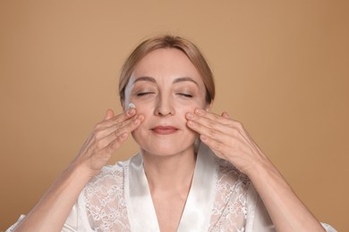 Beautiful woman with cream on face doing self massage against beige background