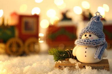 Photo of Cute decorative snowman and toy train on artificial snow against blurred lights, selective focus