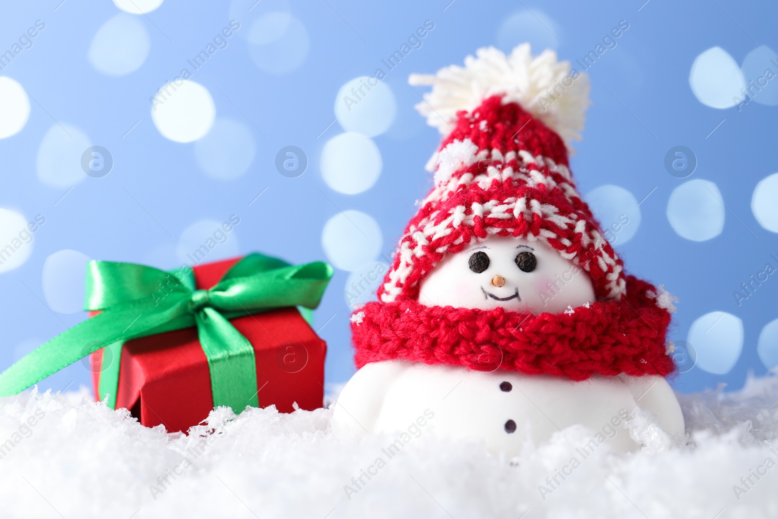 Photo of Cute decorative snowman and gift box on artificial snow against light blue background with blurred lights, closeup