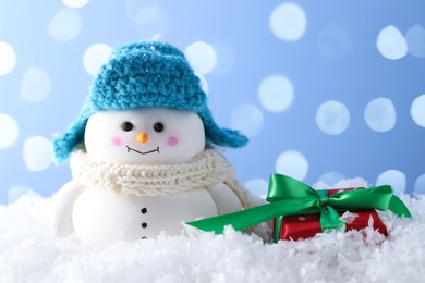 Photo of Cute decorative snowman and gift box on artificial snow against light blue background with blurred lights, closeup