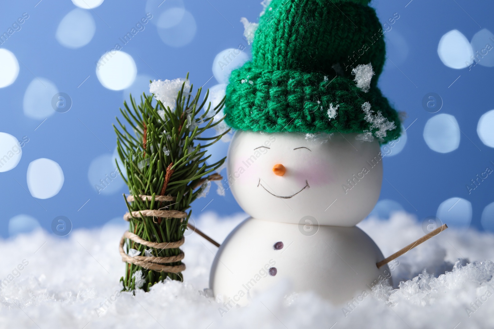 Photo of Cute decorative snowman and fir tree branches on artificial snow against light blue background with blurred lights, closeup