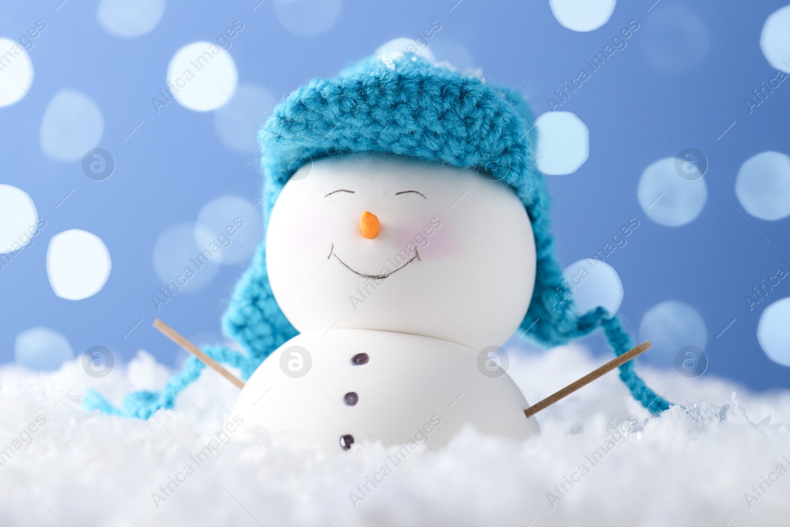 Photo of Cute decorative snowman on artificial snow against light blue background with blurred lights, closeup