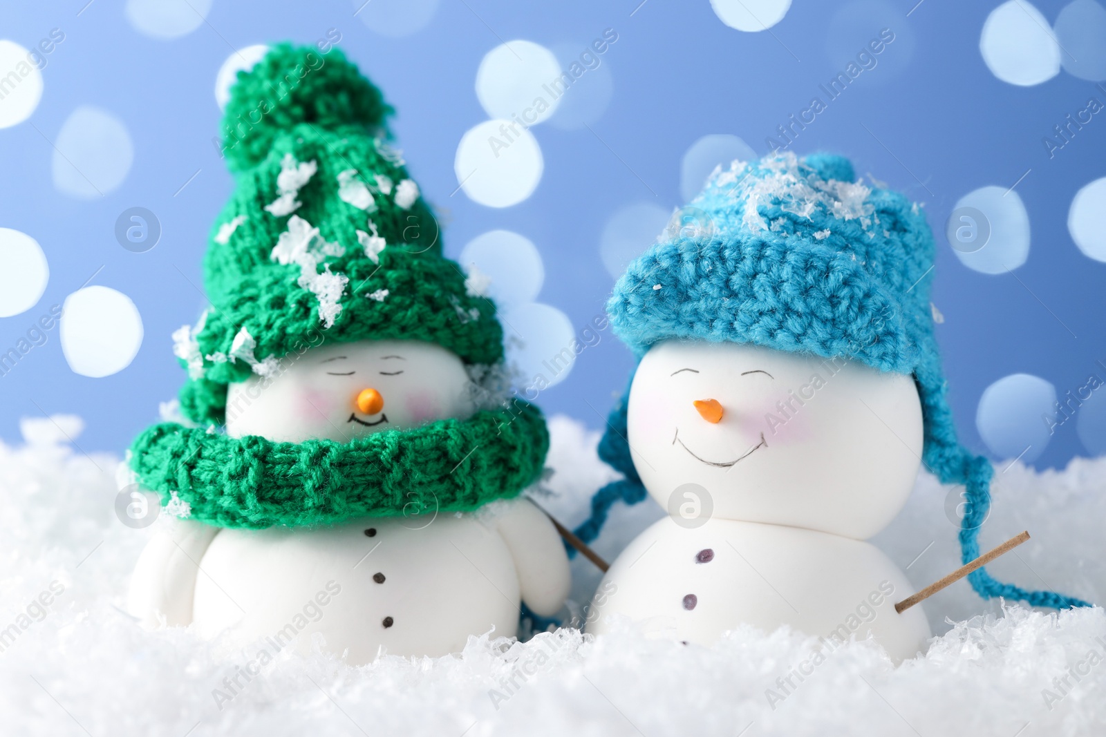 Photo of Cute decorative snowmen on artificial snow against light blue background with blurred lights, closeup