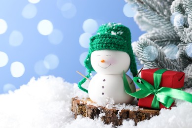 Photo of Cute decorative snowman, gift box and fir tree branches on artificial snow against light blue background with blurred lights, closeup