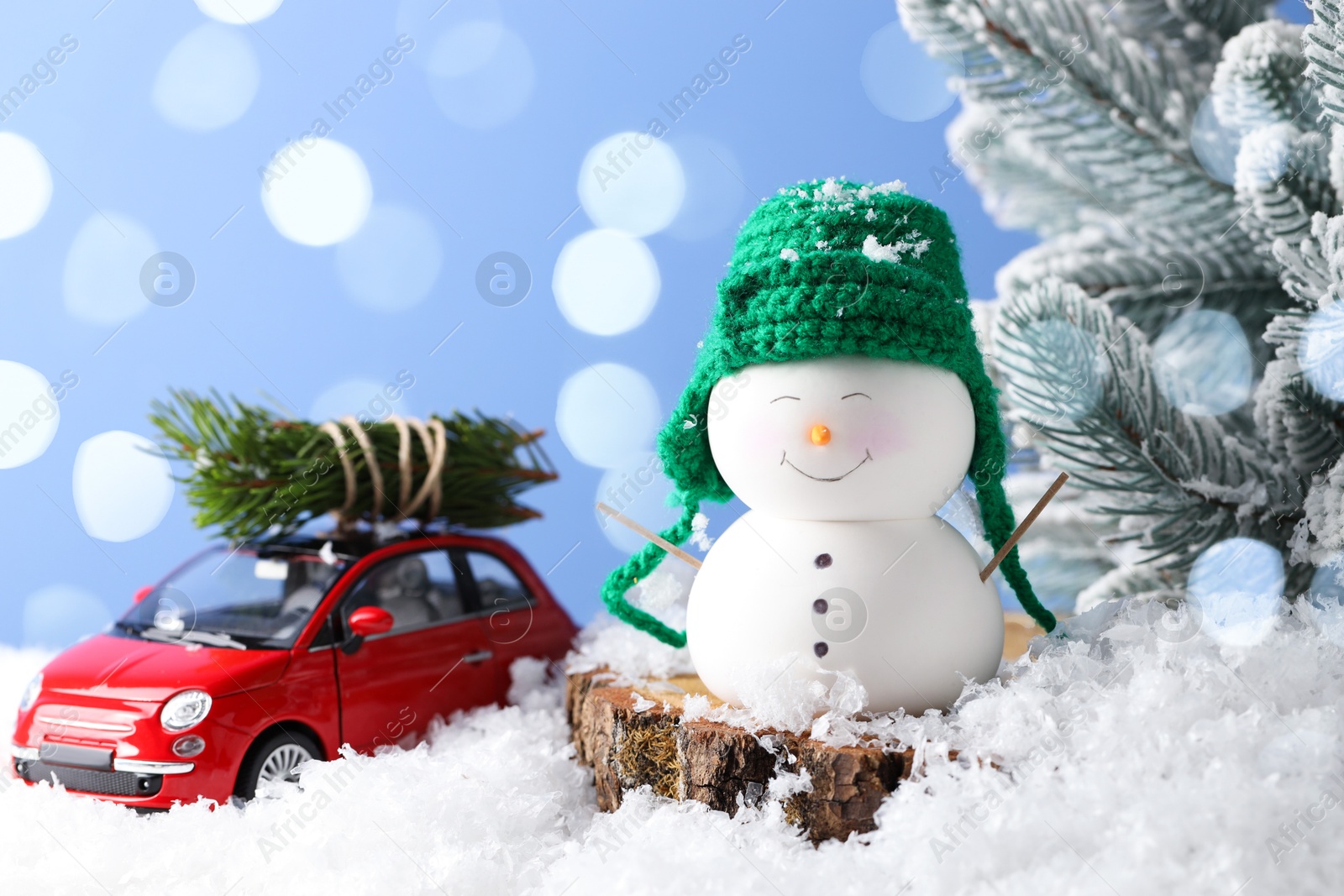 Photo of Cute decorative snowman, toy car and fir tree branches on artificial snow against light blue background, closeup