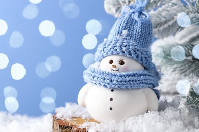 Photo of Cute decorative snowman and fir tree branches on artificial snow against light blue background with blurred lights, closeup. Space for text