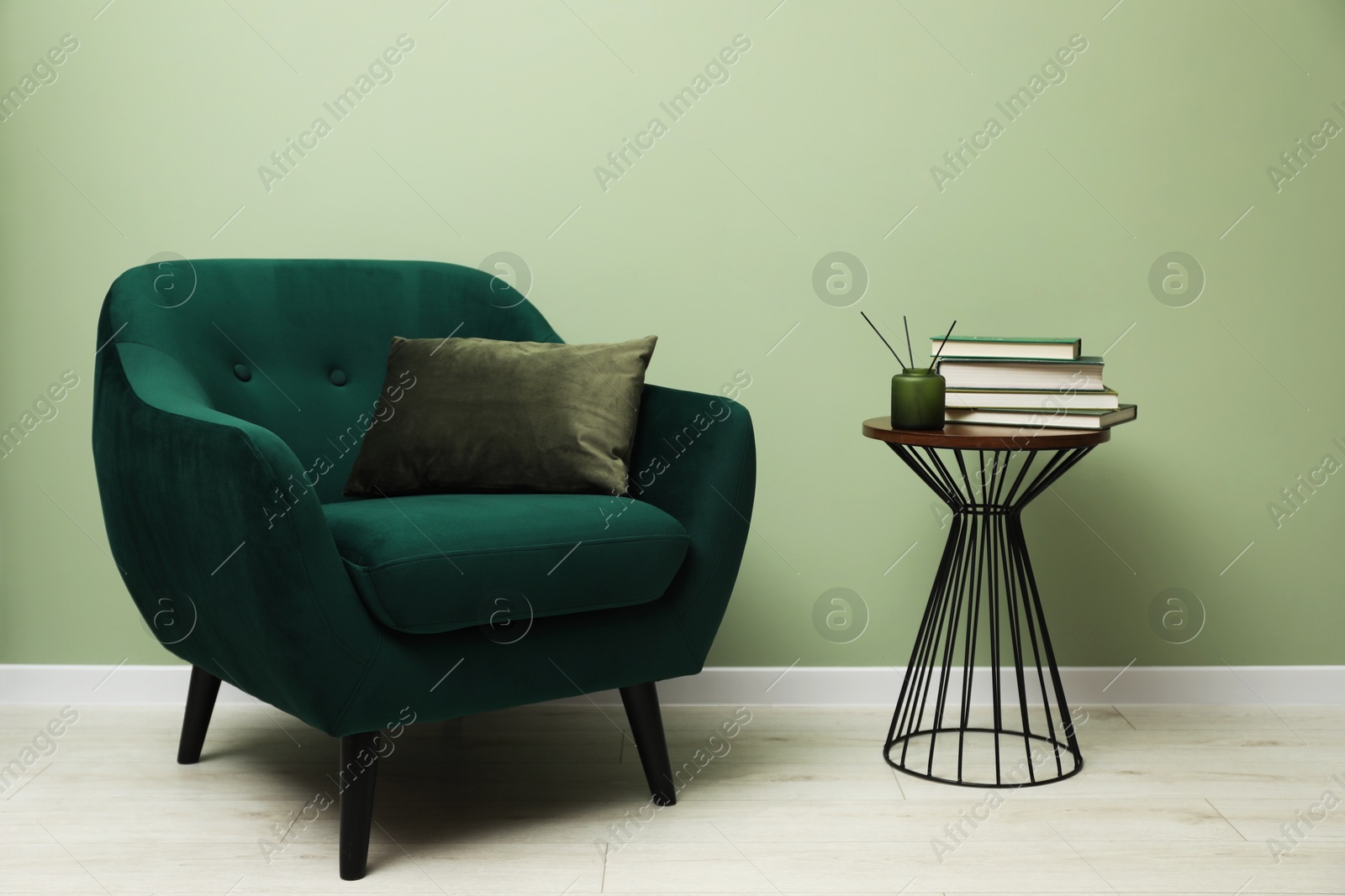 Photo of Stylish green armchair with pillow, books and aromatic reed air freshener on side table near olive wall