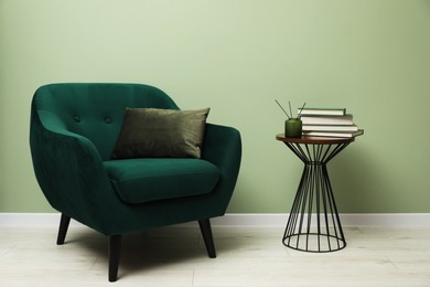 Photo of Stylish green armchair with pillow, books and aromatic reed air freshener on side table near olive wall