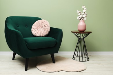 Photo of Stylish green armchair with pink pillow and cotton flowers in vase on side table near olive wall