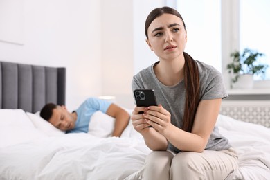 Photo of Frustrated woman with husband's smartphone while he sleeping in bedroom, selective focus