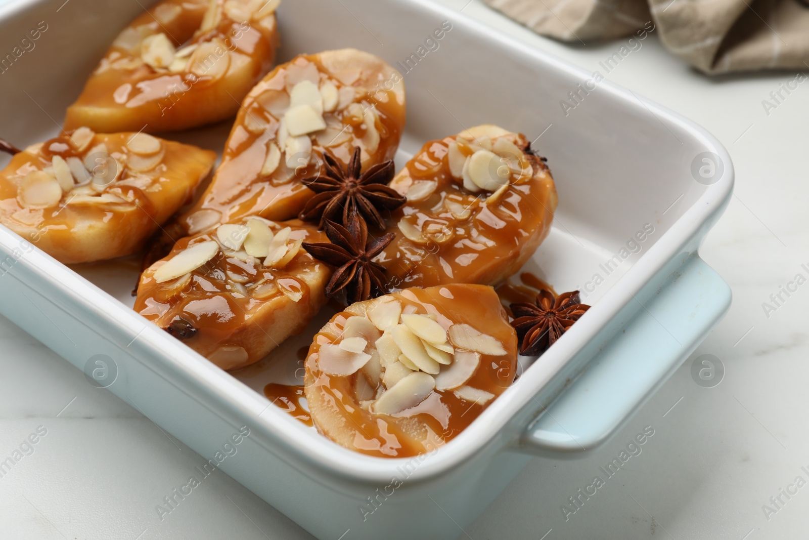 Photo of Delicious pears with caramel sauce, almond flakes and anise stars in baking dish on white marble table, closeup