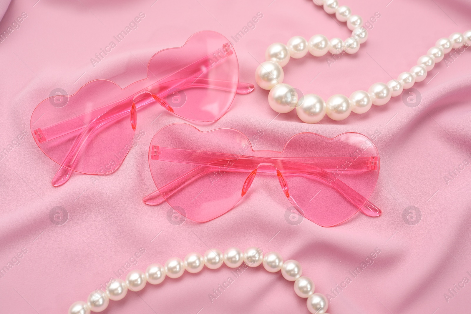 Photo of Stylish heart shaped sunglasses and pearl jewelry on pink fabric