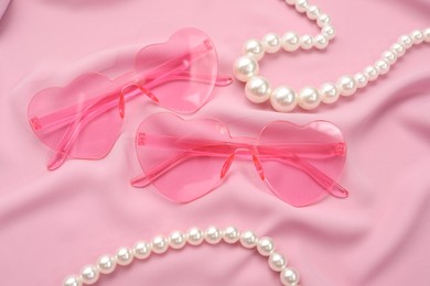 Stylish heart shaped sunglasses and pearl jewelry on pink fabric