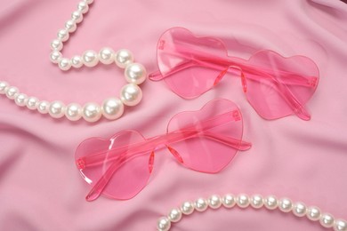 Stylish heart shaped sunglasses and pearl jewelry on pink fabric